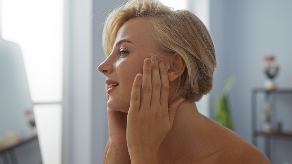 Side profile of a blonde woman with short hair gently touching her jawline and cheek in a softly lit bright interior