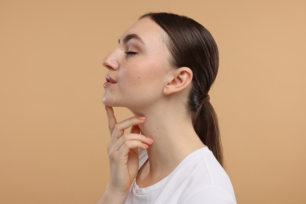 Side profile of a brunette woman with a low ponytail, eyes closed, touching her jawline against a warm beige background
