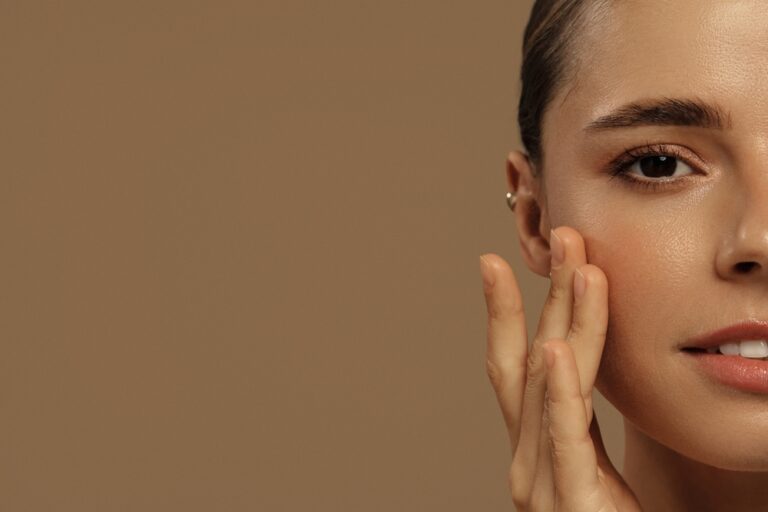 Close-up of a woman with smooth, glowing skin gently touching her cheek against a warm tan background